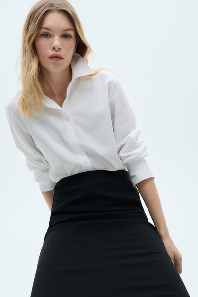 Chemisier blanc à manches longues de MANGO, porté avec une jupe noire unie.
