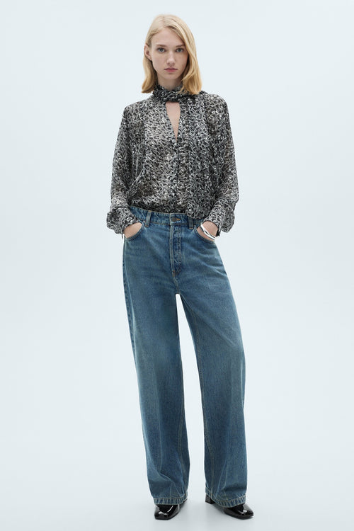 Blouse à manches longues grise de MANGO, avec un motif abstrait, portée avec un jean bleu ample et des chaussures noires vernies.
