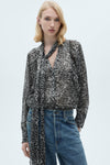 Grijze blouse met lange mouwen van MANGO, met gespikkeld patroon en strikdetail bij de hals, gedragen met een spijkerbroek.