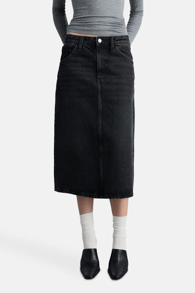 Zwarte midi-spijkerrok van MANGO, van achteren gezien, met zakken en een split aan de achterkant, gedragen met een grijze top en muiltjes.