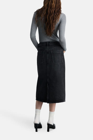 Jupe midi en denim noir par MANGO, vue de dos, avec poches et fente arrière, portée avec un haut gris et des mules.