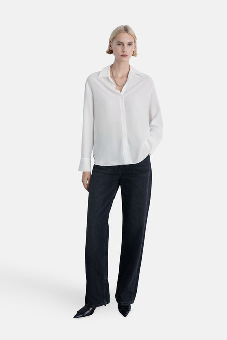 Chemise à manches longues blanche de MANGO, portée avec un pantalon foncé et des chaussures à talons.