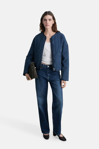 Korte spijkerjas in middinblauw van MANGO, gedragen met een witte tanktop en een blauwe spijkerbroek, met een bruine handtas vast.