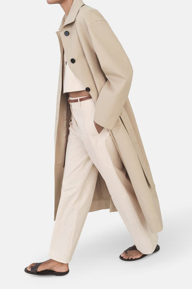 Pantalon écru de MANGO, porté avec une ceinture marron, un top court et un trench beige.