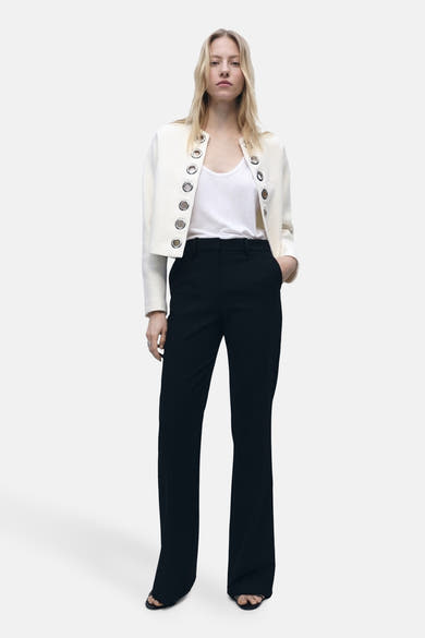 Blazer blanc de MANGO avec des œillets métalliques décoratifs, porté sur un débardeur blanc et un pantalon noir.