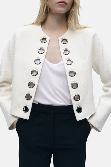 Blazer blanc de MANGO, orné d'œillets argentés, porté sur un t-shirt blanc et un pantalon noir.