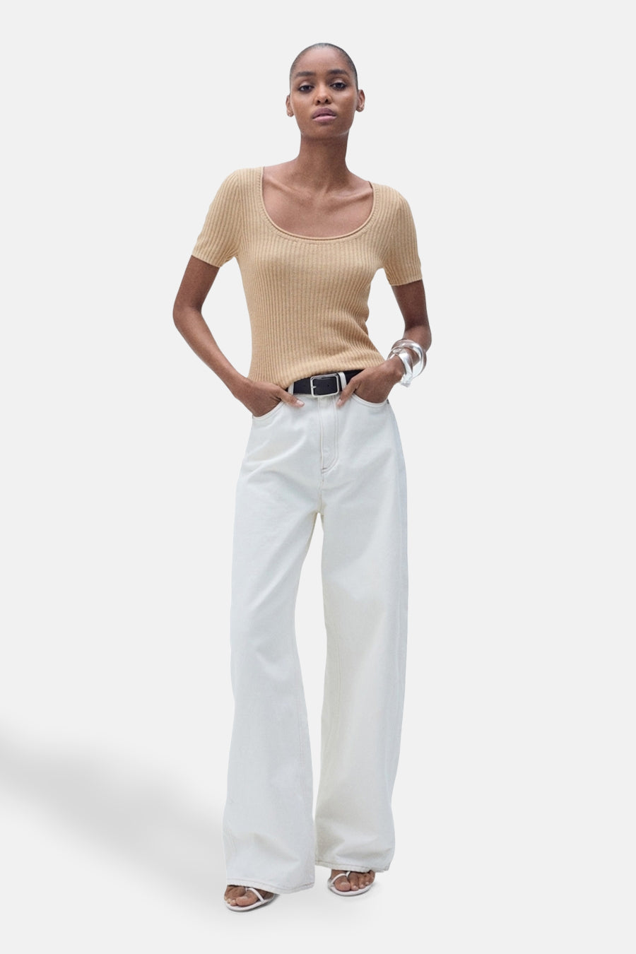 Pull à col rond beige de MANGO, porté avec un pantalon blanc taille haute et une ceinture noire.
