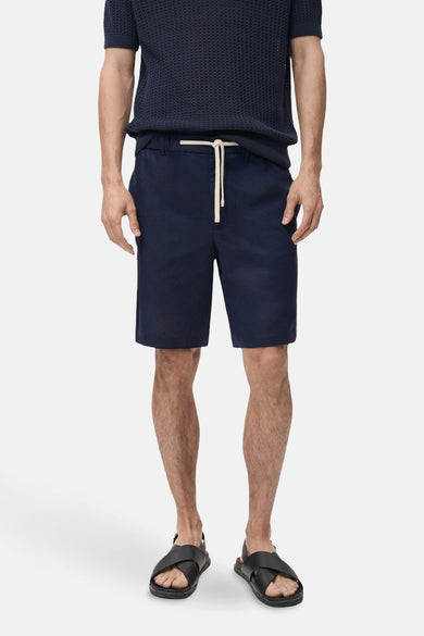 Korte blauwe MANGO-jas, gedragen met een gepoloshirt met textuur en leren sandalen, van achteren gezien.