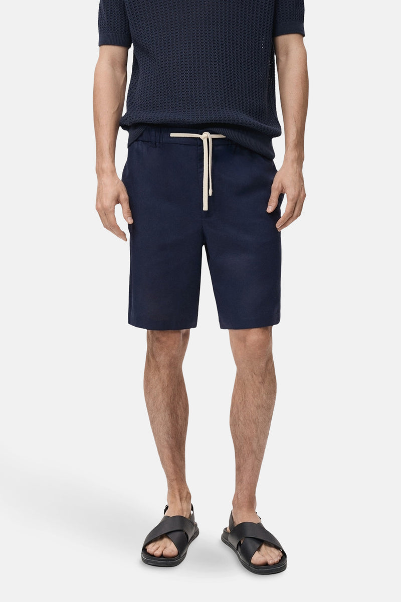 Korte blauwe MANGO-jas, gedragen met een gepoloshirt met textuur en leren sandalen, van achteren gezien.