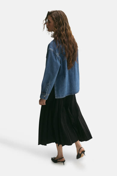 Veste courte en denim bleu moyen de MANGO, portée avec une jupe noire plissée et des talons aiguilles noirs.