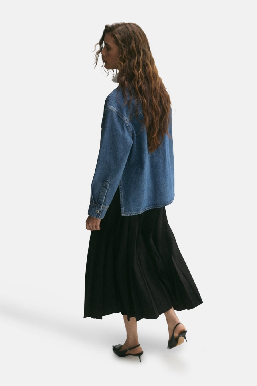 Veste courte en denim bleu moyen de MANGO, portée avec une jupe noire plissée et des talons aiguilles noirs.