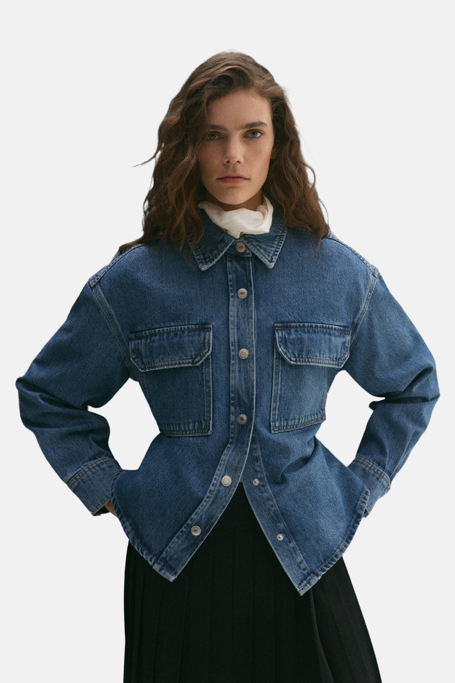 Veste courte en denim bleu moyen de MANGO, avec poches poitrine et coupe ajustée, portée sur une jupe noire plissée.
