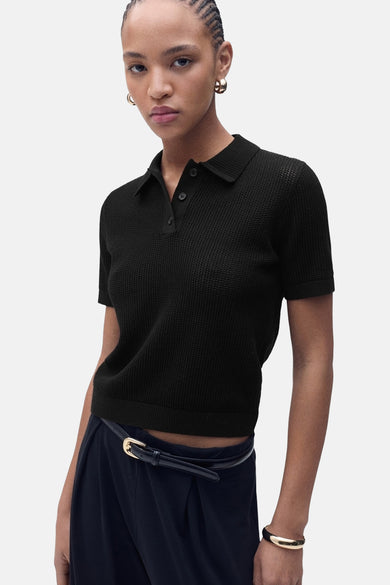 Polo noir à manches courtes de MANGO, avec texture ajourée, porté avec un pantalon taille haute et une ceinture fine.