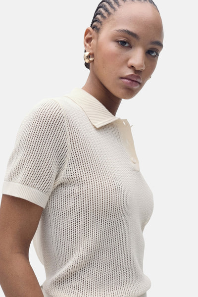 Polo beige à manches courtes de MANGO, en maille ajourée, porté avec des boucles d'oreilles dorées.