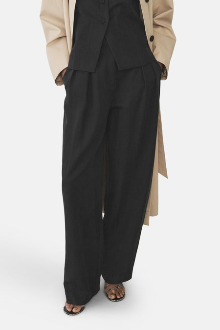 Zwarte pantalon met plooien van MANGO, losse pasvorm, gedragen met een zwarte top en een beige trenchcoat.