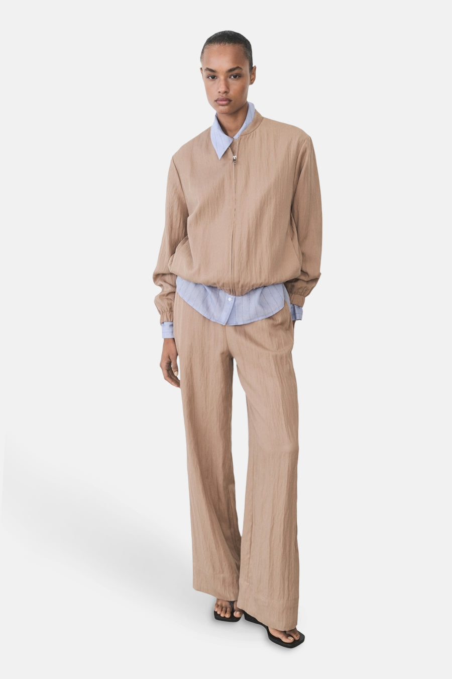 Veste courte brune de MANGO, portée sur une chemise bleue à rayures, avec un pantalon assorti et des sandales noires.
