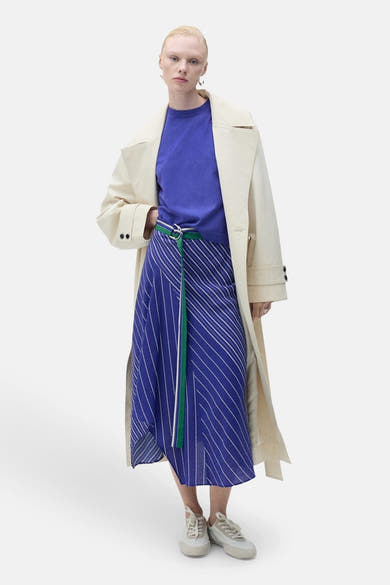 Pull à col rond bleu de MANGO, porté avec une jupe à rayures et un trench beige.