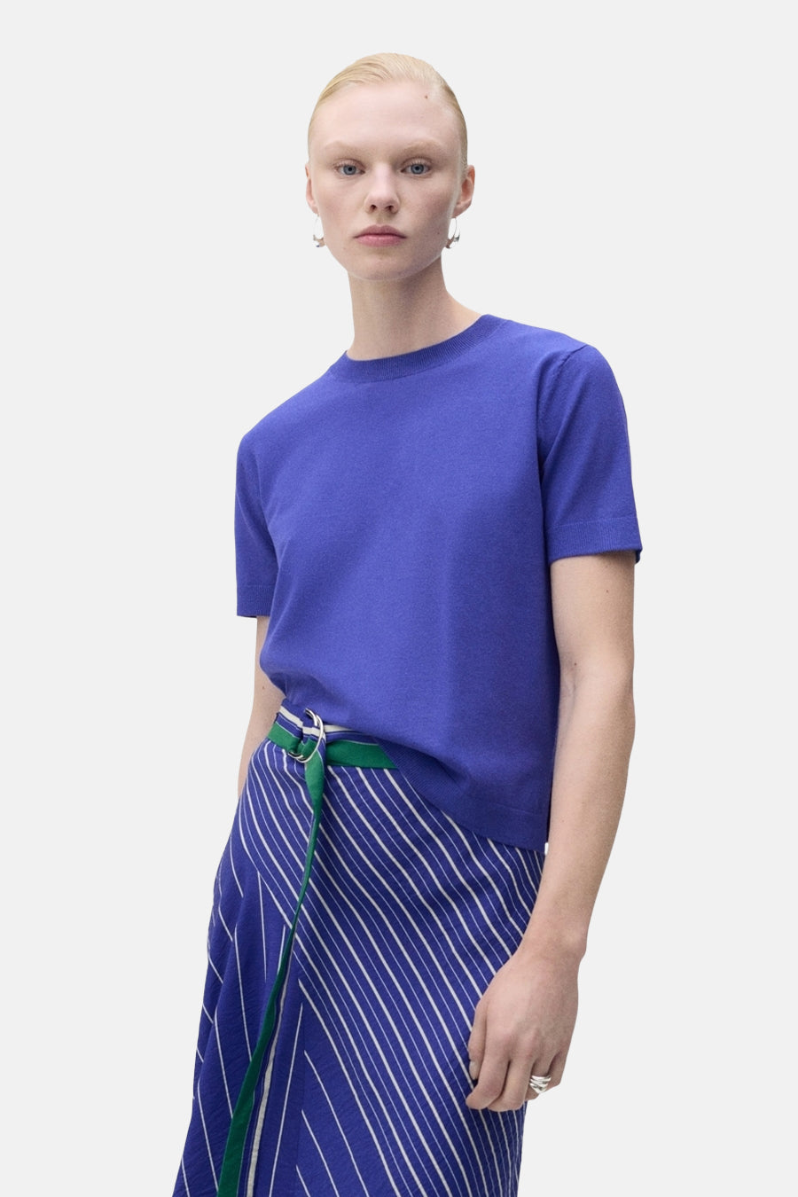 Pull à col rond bleu de MANGO, porté avec une jupe à rayures bleues et blanches et une ceinture verte.
