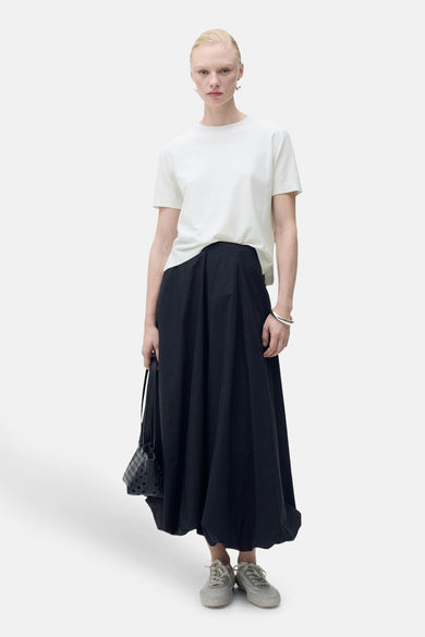 Pull à col rond blanc de MANGO, porté avec une jupe noire ample, un bracelet argenté et un petit sac.