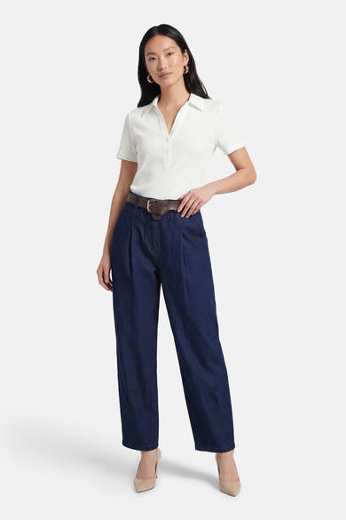 Pantalon en denim bleu foncé, de Terra di Siena, avec une ceinture marron et des plis, porté avec un polo blanc et des escarpins nude.

