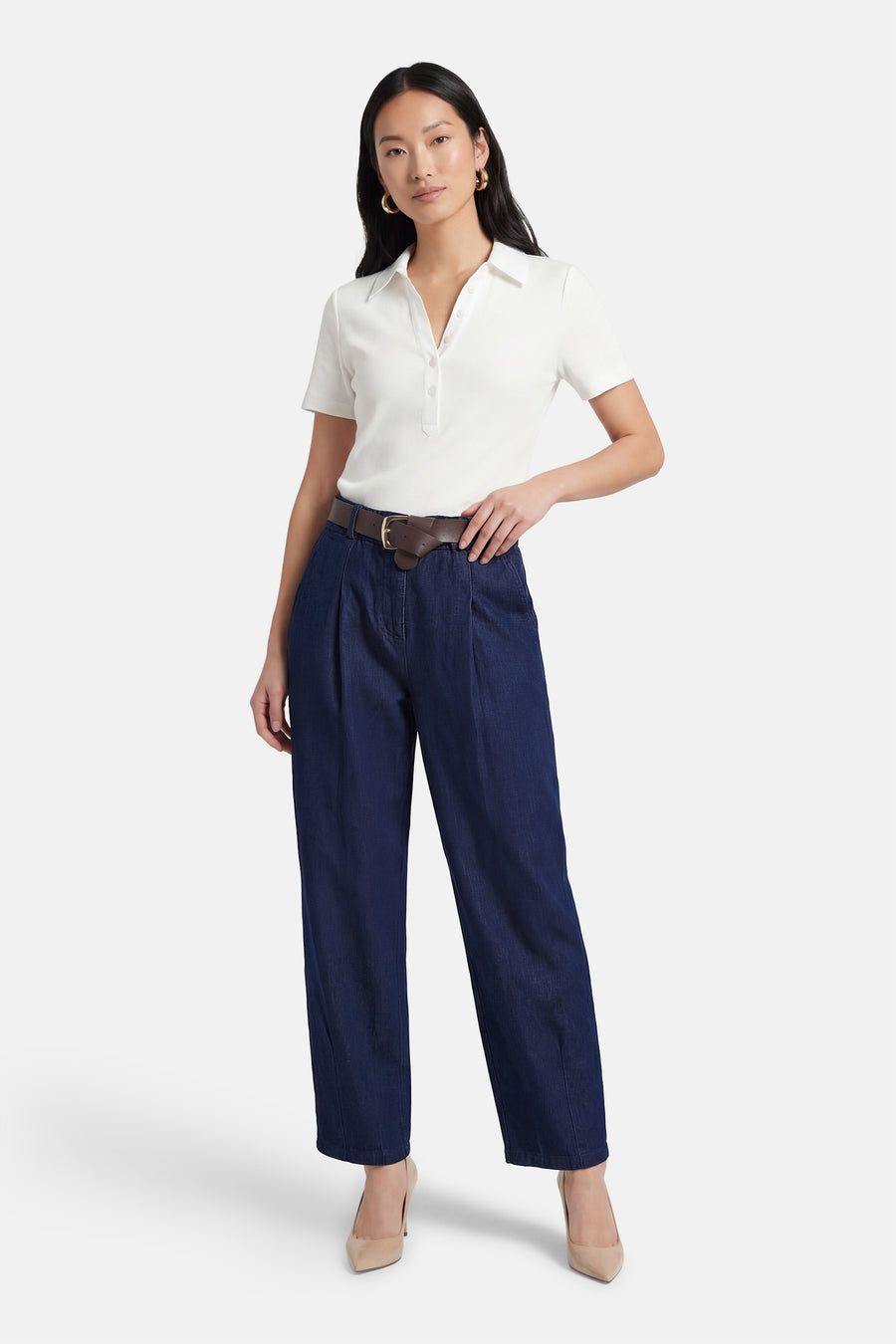 Pantalon en denim bleu foncé, de Terra di Siena, avec une ceinture marron et des plis, porté avec un polo blanc et des escarpins nude.

