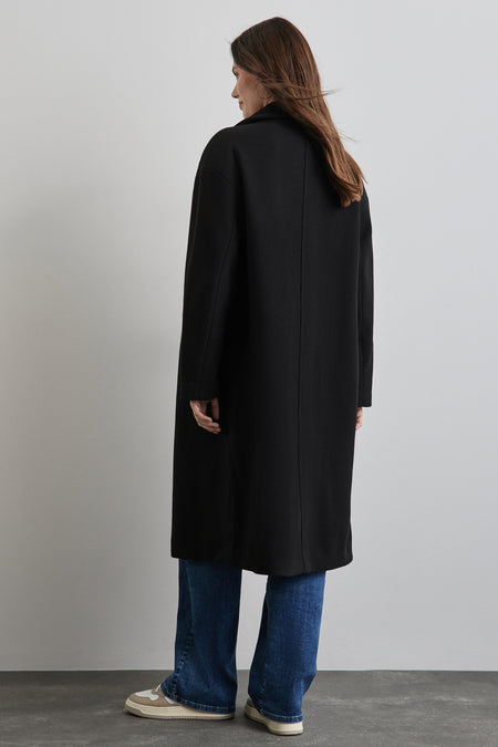 Lange zwarte jas van STREET ONE, gedragen met een blauwe jeans en beige sportschoenen, van achteren gezien.