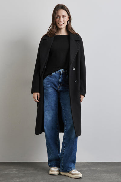 Veste longue noire de Street One, portée avec un pull noir, un jean bleu et des baskets beiges.

