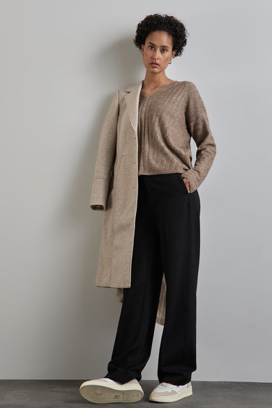 Pull à col en V beige de Street One, avec texture côtelée, porté avec un pantalon noir et un manteau beige posé sur l'épaule.
