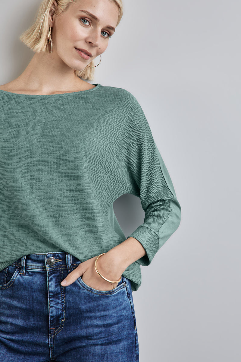 T-shirt vert de STREET ONE, manches longues amples, porté avec un jean bleu et un bracelet doré.
