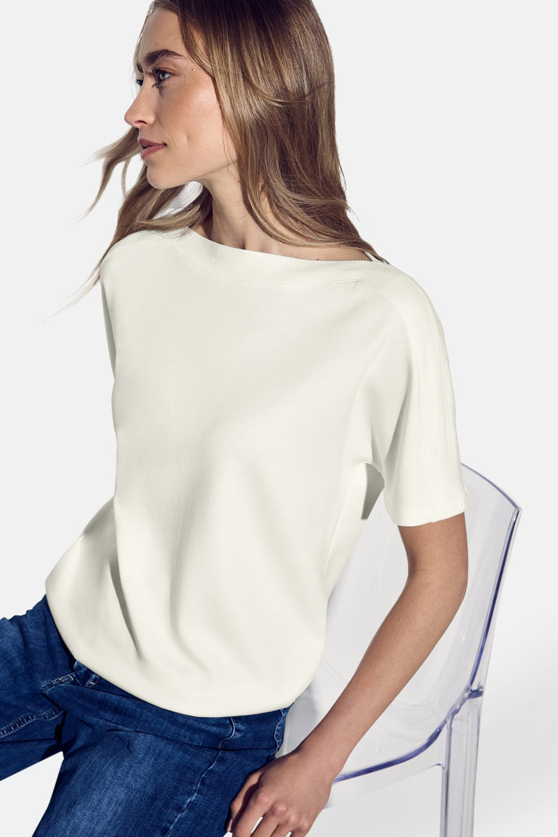 Top met korte mouwen wit van Street One, gedragen met een blauwe jeans en zittend op een transparante stoel.