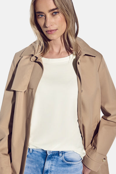 Top street one korte mouwen in ecru, gedragen met een beige trenchcoat en een blauwe jeans.