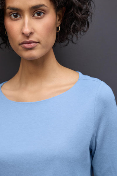 T-shirt bleu à manches longues de STREET ONE, porté avec des boucles d'oreilles dorées et cheveux bruns bouclés.
