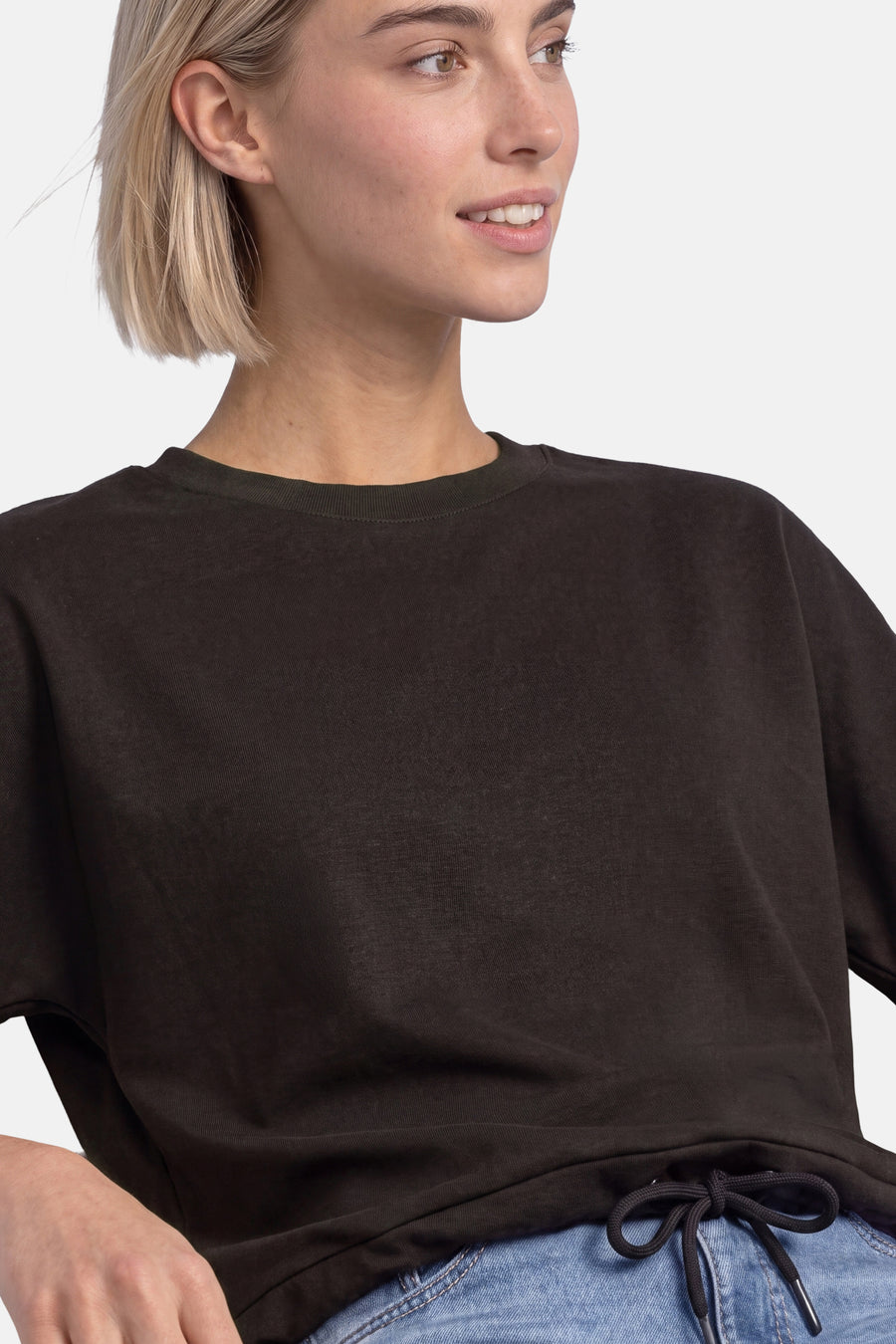 Brown crewneck sweatshirt with ribbed neckline and relaxed fit, worn with light blue jeans and tied hem detail, side view close-up