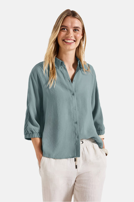 Blauwe blouse met lange mouwen van Street One, gedragen met een witte broek en gouden sieraden.