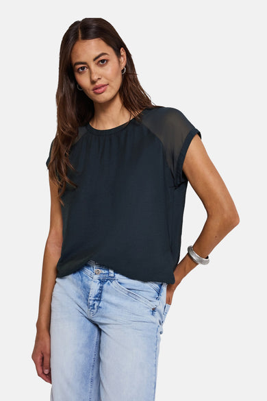 Blauwe blouse met korte mouwen van STREET ONE, met doorschijnende inzetten op de schouders, gedragen met een verbleekte spijkerbroek en een zilveren armband.