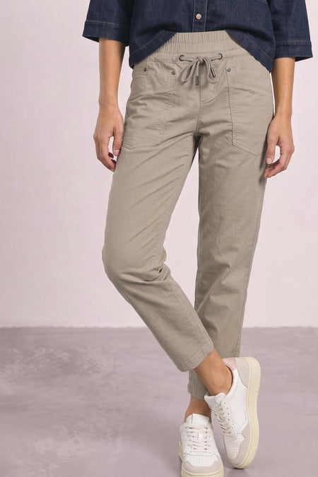 Beige cotton pants with elastic waist and drawstring, featuring front pockets and cuffed ankles, worn with a navy shirt and white sneakers in a lifestyle pose.