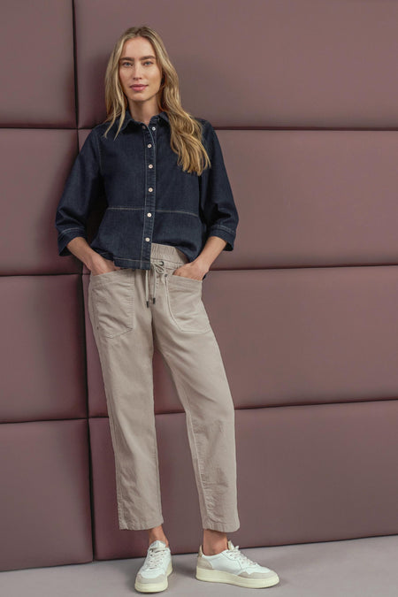 Beige cotton pants with drawstring waist and side pockets, worn with dark denim shirt and white sneakers, full-length lifestyle view