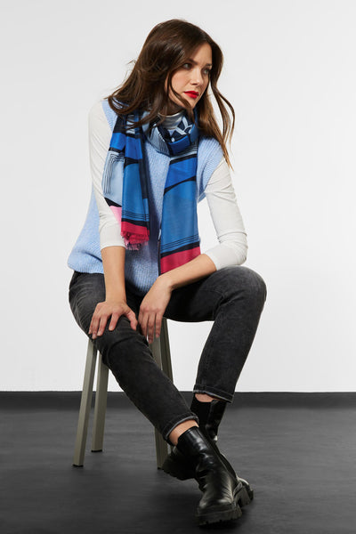 Blue patterned foulard with geometric stripes and fringed edges, worn by model in lifestyle pose with light blue vest and dark jeans.