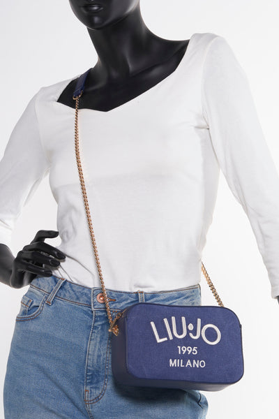 Sac à bandoulière bleu de LIU JO, avec une bandoulière chaîne dorée, porté avec un t-shirt blanc et un jean bleu clair.
