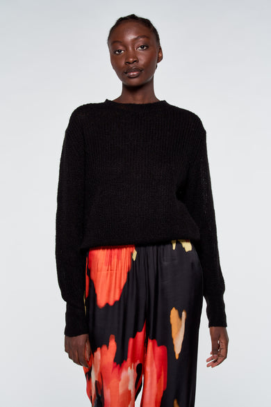 Pull noir par ASTRID Black Label, porté avec un pantalon à motif floral rouge et jaune.