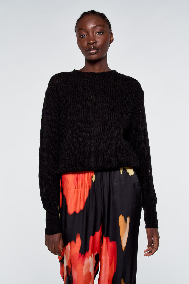 Pull noir par ASTRID Black Label, porté avec un pantalon à motif floral rouge et jaune.