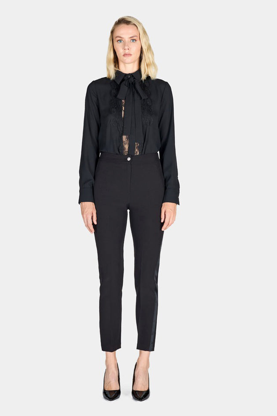 Pantalon coloré noir ARTIGLI à coupe cigarette, avec une bande latérale satinée et porté avec un chemisier noir à dentelle.

