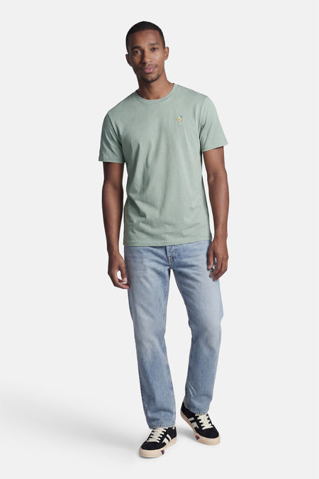 Groen T-shirt met korte mouwen van STROM met cocktailborduurwerk op de borst, gedragen door een man in een spijkerbroek.