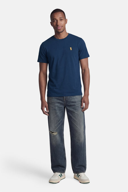 Blauw T-shirt met korte mouwen van STROM, met een oranje embleem op de borst, gedragen met een spijkerbroek en witgroene sneakers.