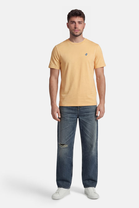 Gele T-shirt met korte mouwen van STROM met een klein blauw "S"-logo, gedragen met een gescheurde spijkerbroek en witte sneakers.