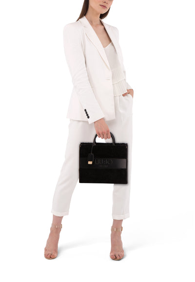 Sac à bandoulière noir en fausse fourrure de LIU JO, porté avec un tailleur pantalon blanc et des sandales transparentes.