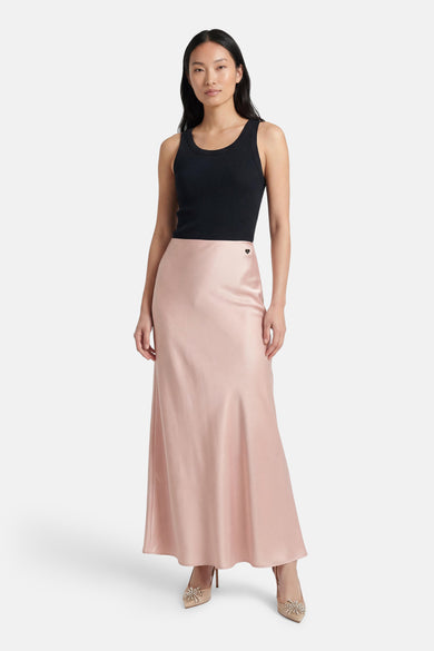 Jupe longue rose en satin d'Amélie et Amélie, avec un petit logo en cœur et portée avec un débardeur noir et des escarpins beiges.
