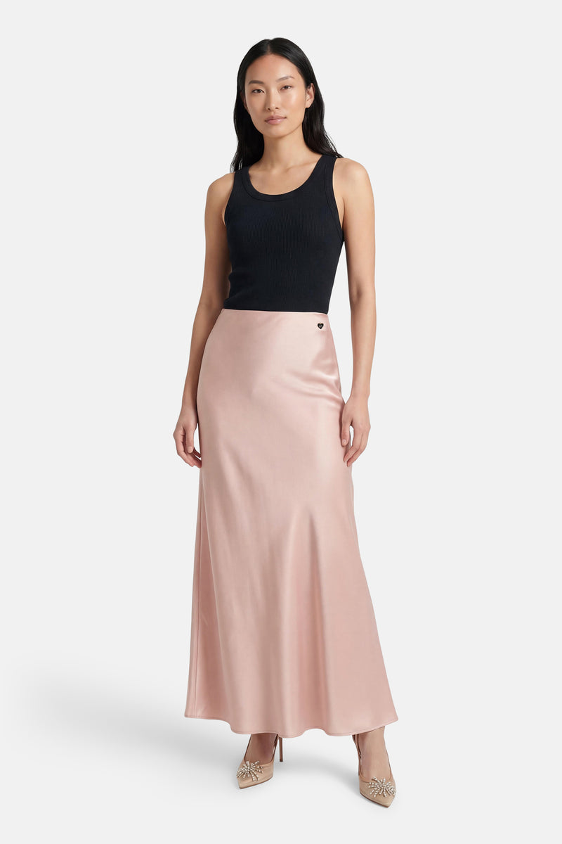 Jupe longue rose en satin d'Amélie et Amélie, avec un petit logo en cœur et portée avec un débardeur noir et des escarpins beiges.
