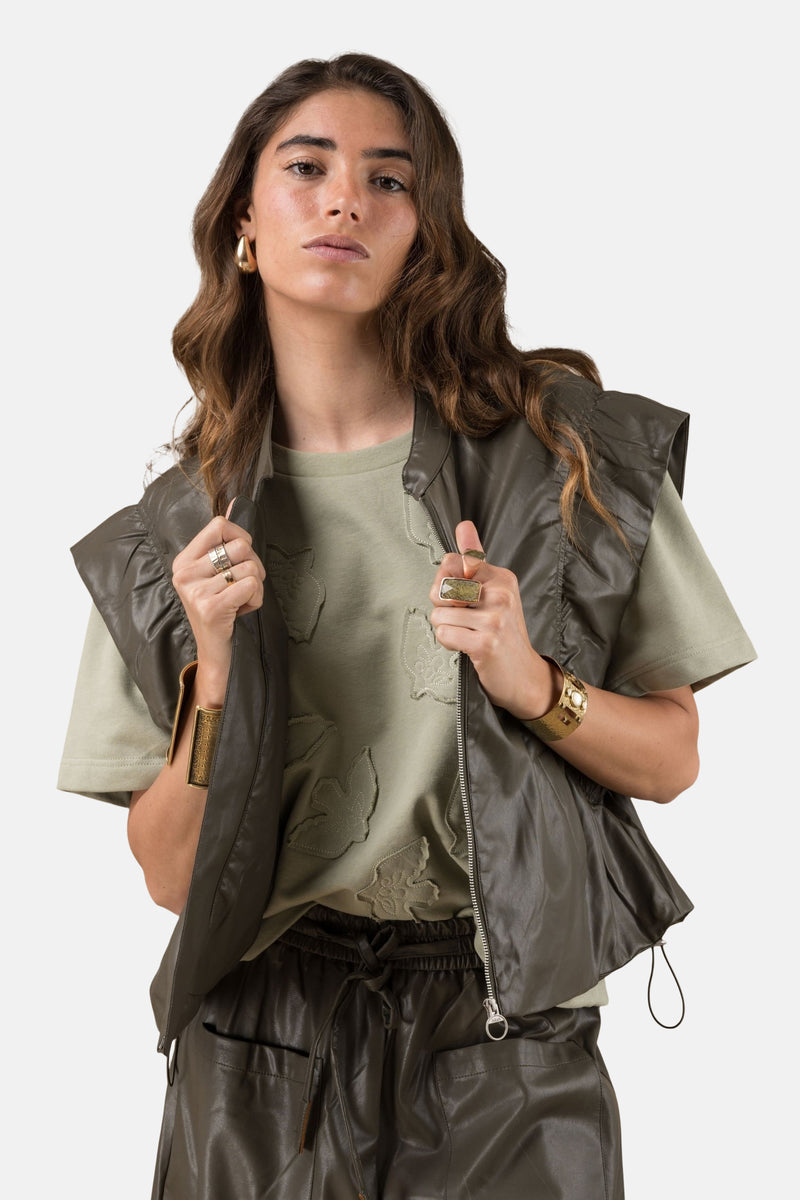 Gilet kaki, de ADORE, avec fermeture éclair, porté sur un t-shirt vert, et des bijoux dorés.
