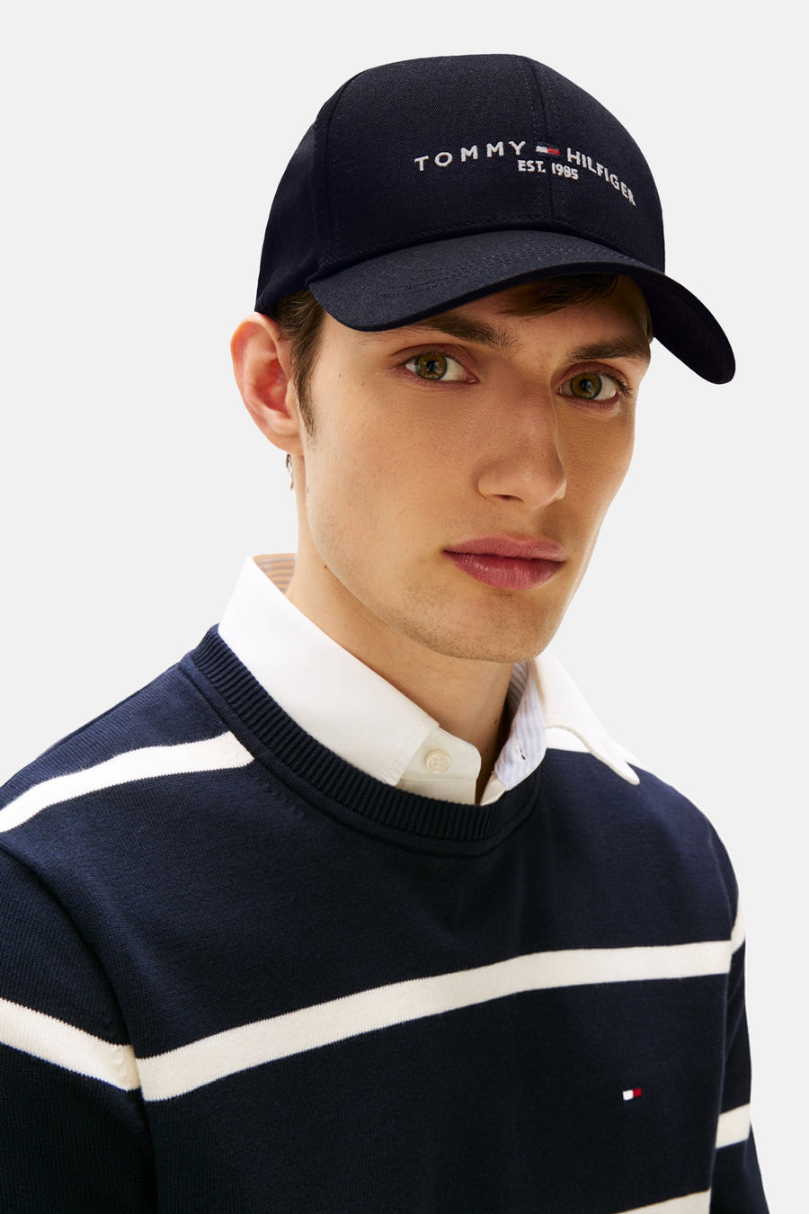 Casquette bleue de TOMMY JEANS, avec logo brodé, portée avec un pull rayé et une chemise blanche.
